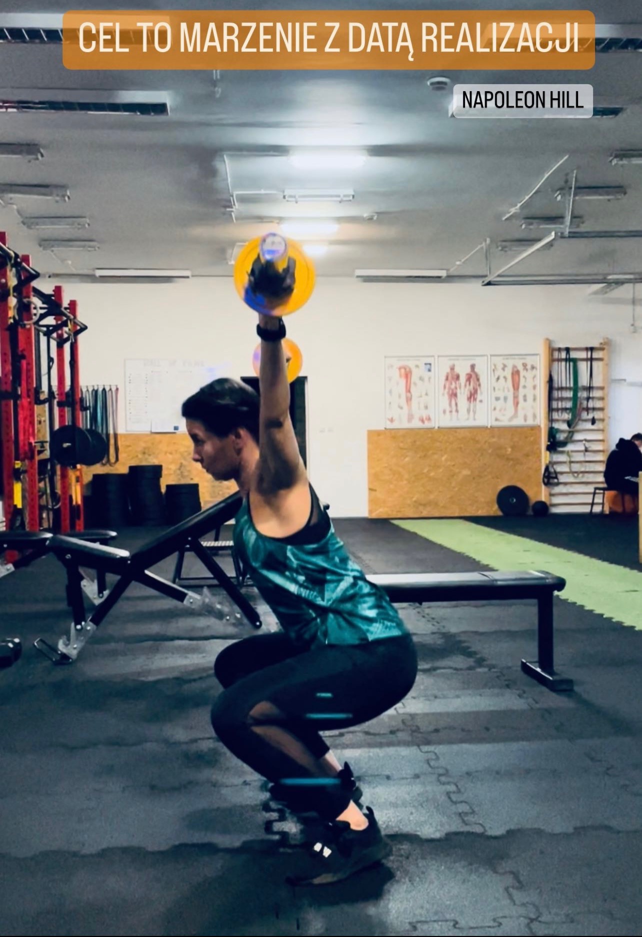 Kobieta w sportowym stroju wykonuje przysiad z obciążeniem kettlebell nad głową w siłowni; widoczne plakaty anatomiczne i sprzęt do ćwiczeń w tle.