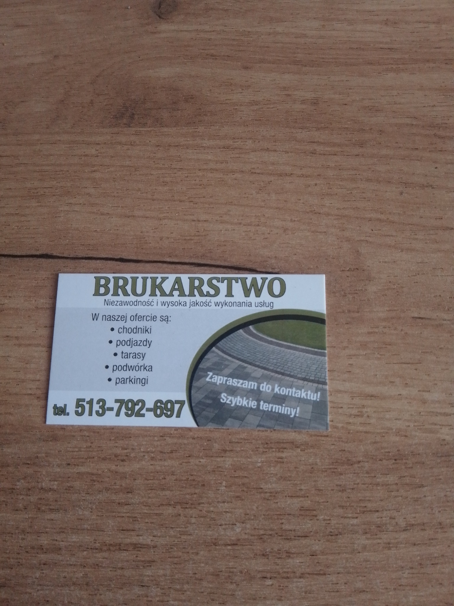 Wizytówka firmy brukarskiej na drewnianym tle. Oferta obejmuje chodniki, podjazdy, tarasy, podwórka i parkingi. Numer telefonu 513-792-697. Zaproszenie do kontaktu i szybkie terminy.