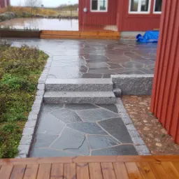 Ścieżka z nieregularnych płyt kamiennych obramowana kostką, prowadząca do domu z czerwonymi ścianami i drewnianym tarasem, widoczna po deszczu.