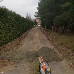 Przygotowanie pod utwardzenie drogi dojazdowej: wyrównany teren, pryzma ziemi, szpadel i przecinarka spalinowa Stihl, w tle szpaler tuj.