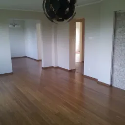 Jasne, przestronne wnętrze z parkietem bambusowym, ozdobnym żyrandolem i widocznymi przejściami do innych pomieszczeń. Ściana po prawej stronie z dekoracyjnym wykończeniem imitującym kamień.