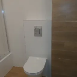 Nowoczesna toaleta podwieszana z chromowanym przyciskiem spłukującym, białe płytki ścienne zestawione z brązowymi płytkami imitującymi drewno na podłodze i ścianie.