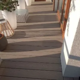 Balkon wyłożony kompozytowymi panelami imitującymi drewno, z białymi donicami z roślinami i rattanowym stolikiem, oświetlony popołudniowym słońcem.