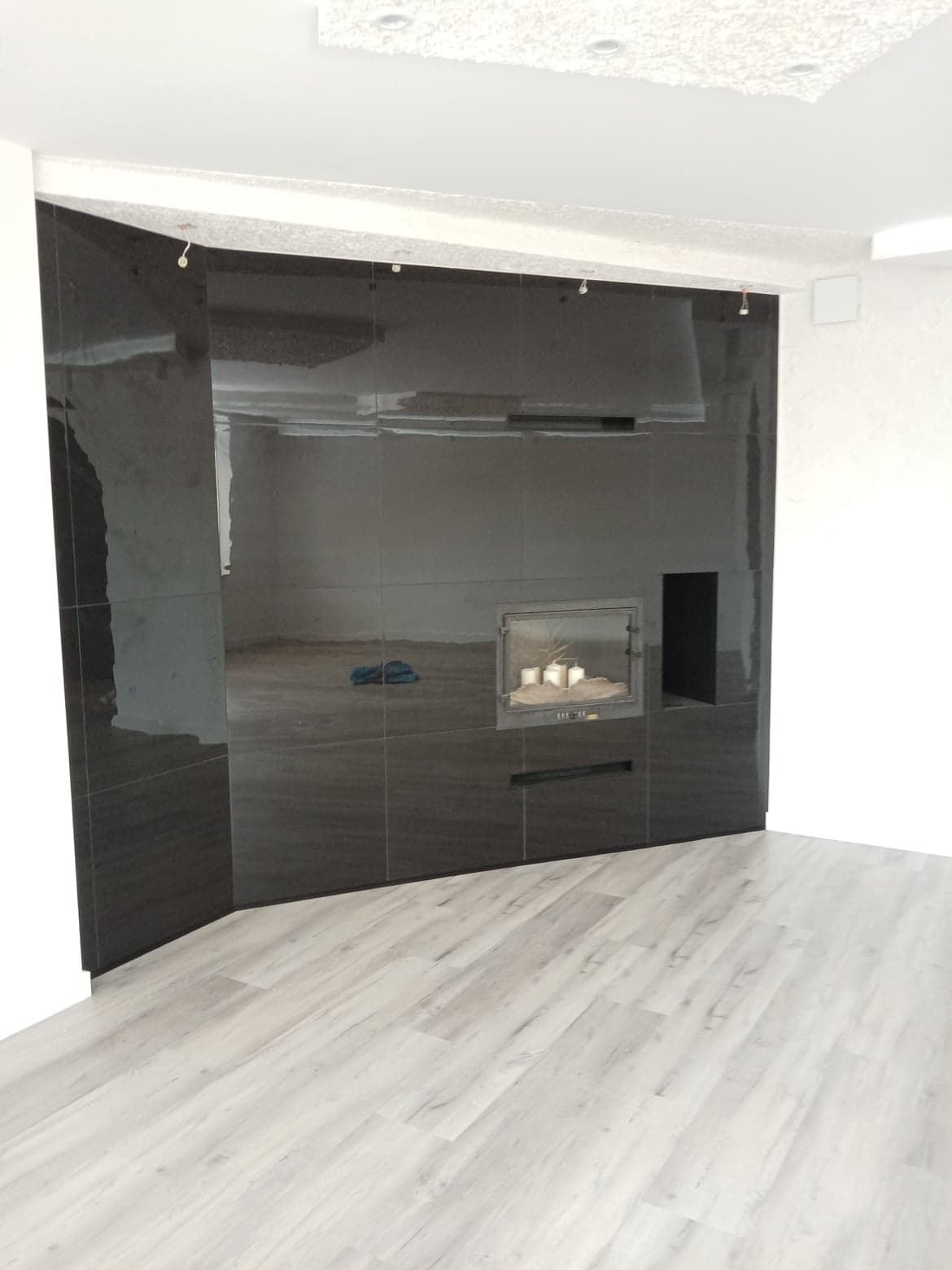 Ciemna ściana wyłożona błyszczącymi kaflami, z wbudowanym kominkiem i jasną podłogą z paneli imitujących drewno.