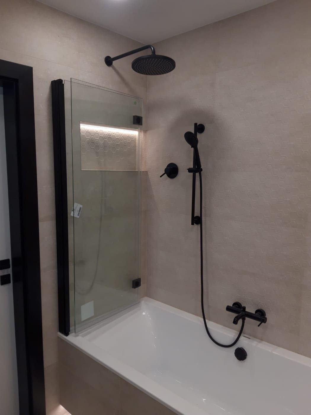 Nowoczesna łazienka z czarną armaturą: deszczownica, bateria wannowa i szklany parawan z czarną ramą nad białą wanną, podświetlana wnęka z mozaiką heksagonalną.