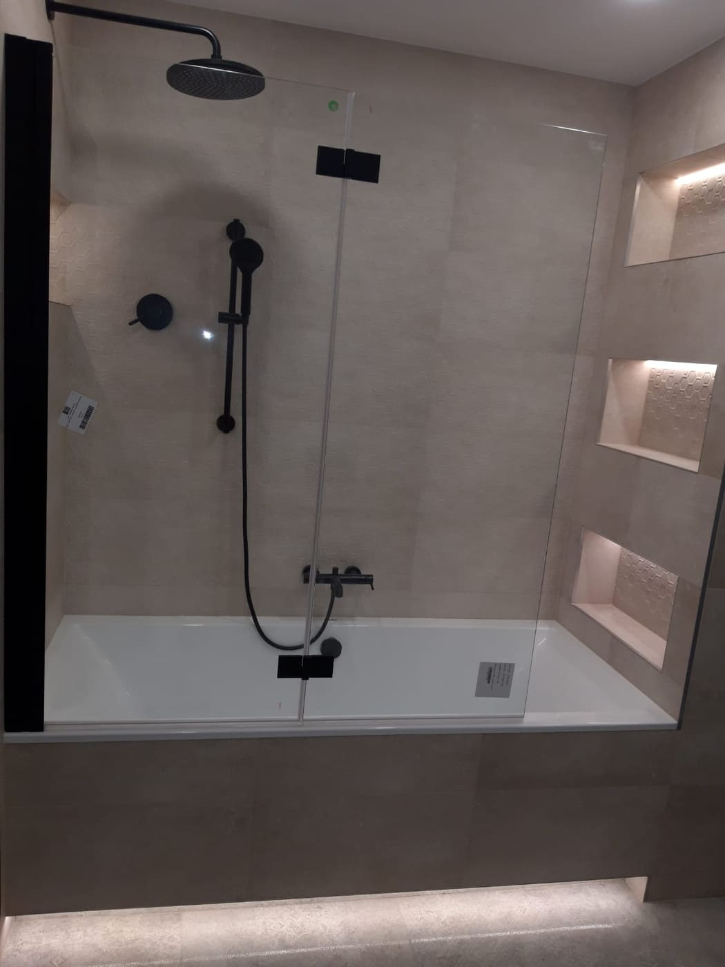 Nowoczesna łazienka z wanną i szklanym parawanem nawannowym z czarnymi elementami, w tle wnęki z podświetlaną mozaiką.