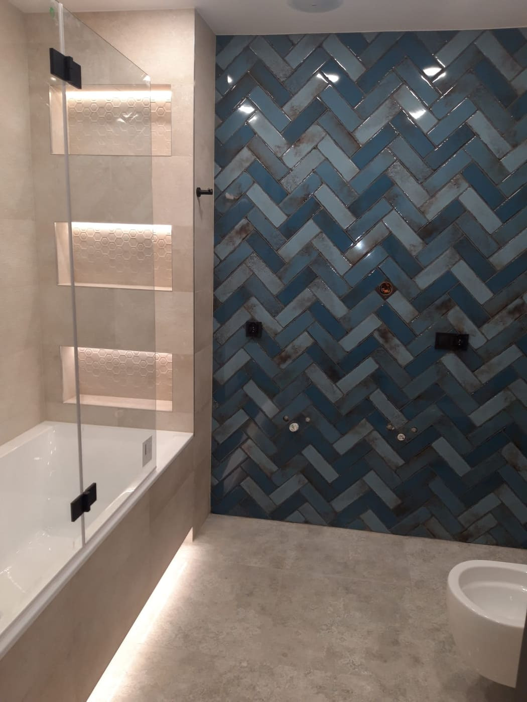 Nowoczesna łazienka z niebieską ścianą z płytek ułożonych w jodełkę, szklany parawan nawannowy z czarnymi okuciami, podświetlane półki w ścianie i minimalistyczna toaleta.