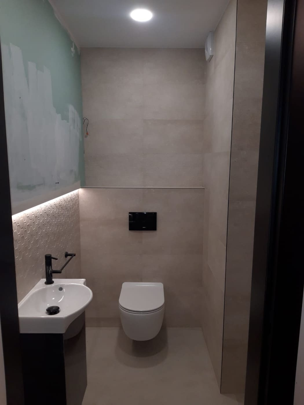 Nowoczesna łazienka w trakcie wykańczania: widoczna umywalka z czarną armaturą, podświetlana mozaika, wisząca toaleta i fragment ściany przygotowanej do malowania.