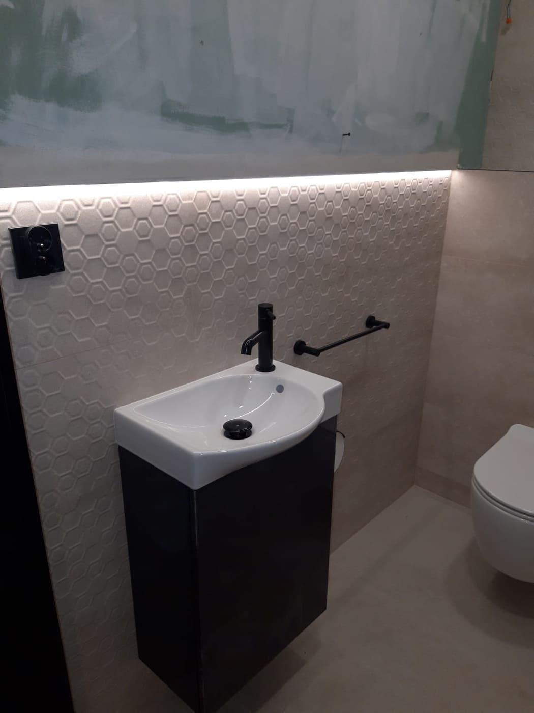 Mała umywalka z czarną szafką i baterią, heksagonalne płytki na ścianie podświetlone ledami, widoczne niedokończone malowanie ściany w górnej części.