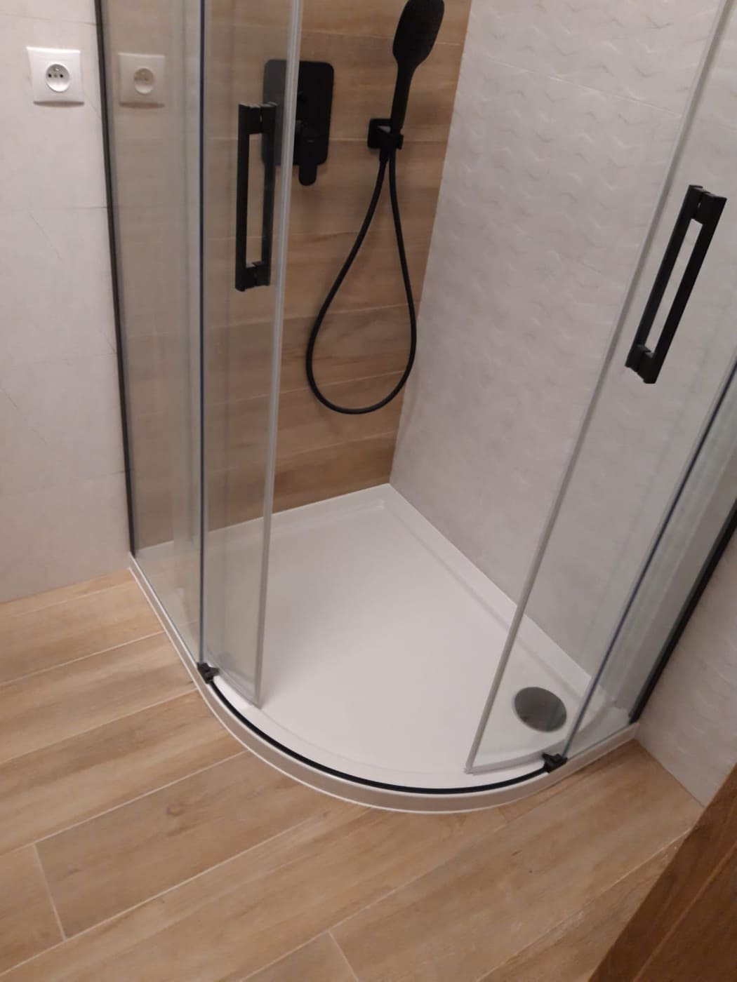 Nowoczesna kabina prysznicowa z czarną armaturą, biały brodzik i drewnopodobne płytki podłogowe, widoczne dwa gniazdka elektryczne na ścianie.