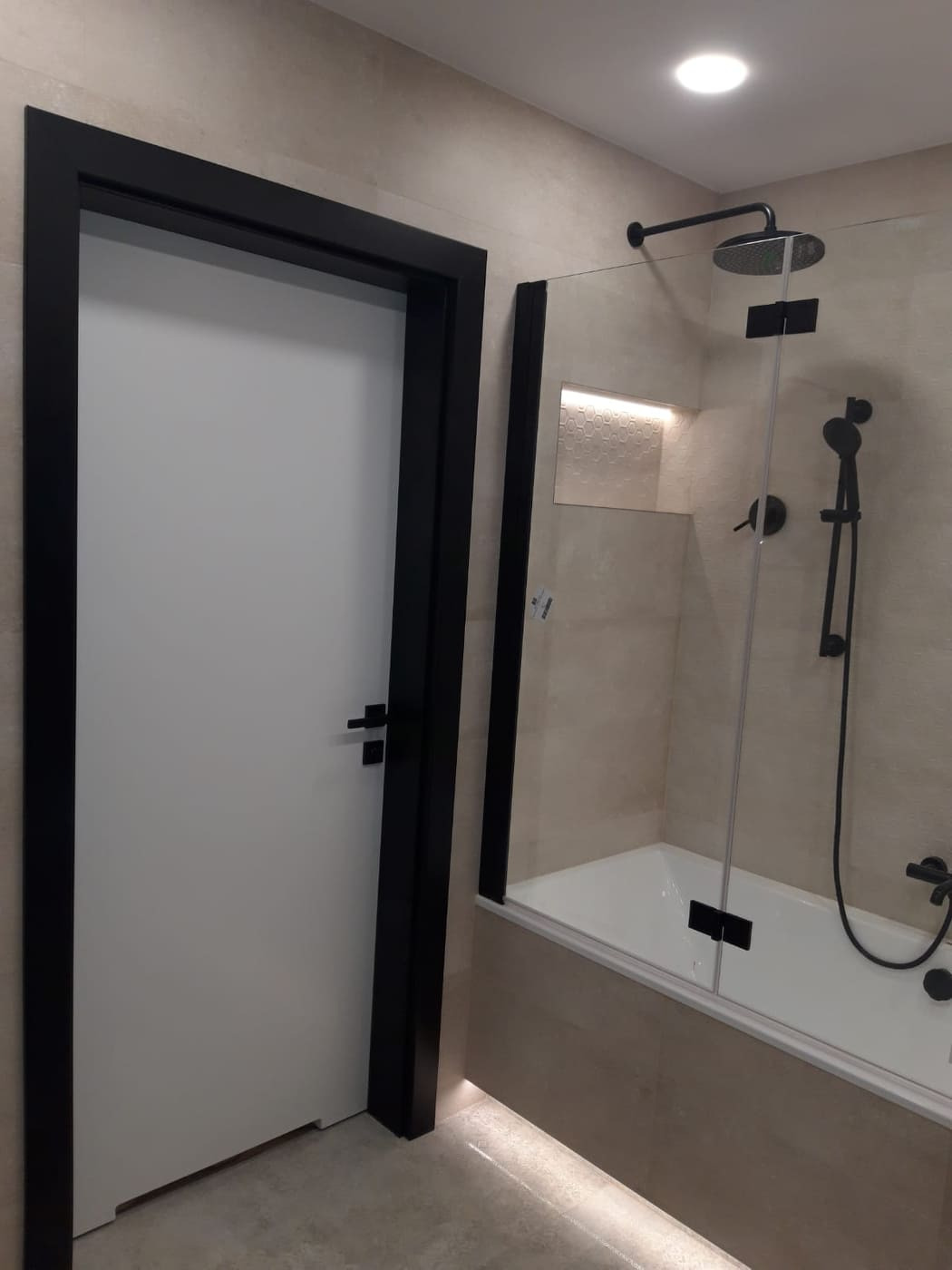 Nowoczesna łazienka z białą wanną z czarną armaturą, szklanym parawanem nawannowym i białą, zamkniętą drzwiami z czarną ościeżnicą i klamką