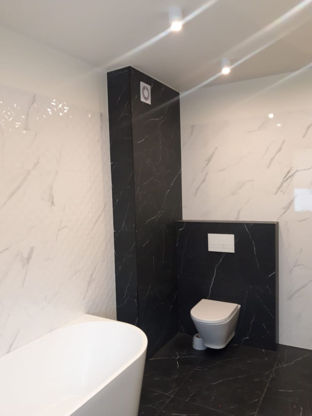 Nowoczesna łazienka z białą wanną, wiszącą toaletą i czarnymi płytkami imitującymi marmur na podłodze i ścianach, z białymi płytkami o strukturze 3D na ścianie.