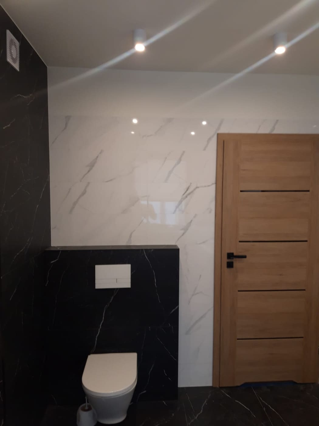 Nowoczesna łazienka z czarno-białymi marmurkowymi płytkami, drewnianymi drzwiami z poziomymi frezowaniami i podwieszaną toaletą z białym przyciskiem spłukującym.