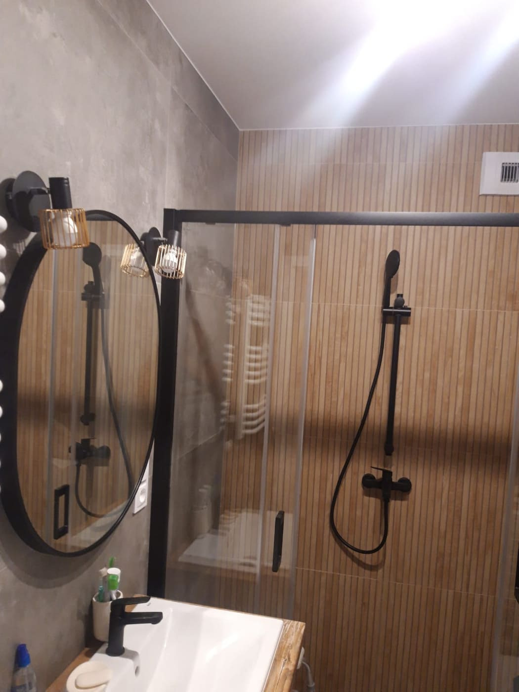 Nowoczesna łazienka z kabiną prysznicową typu walk-in, wykończona płytkami imitującymi drewno, z czarną armaturą i owalnym lustrem z ozdobnym oświetleniem.
