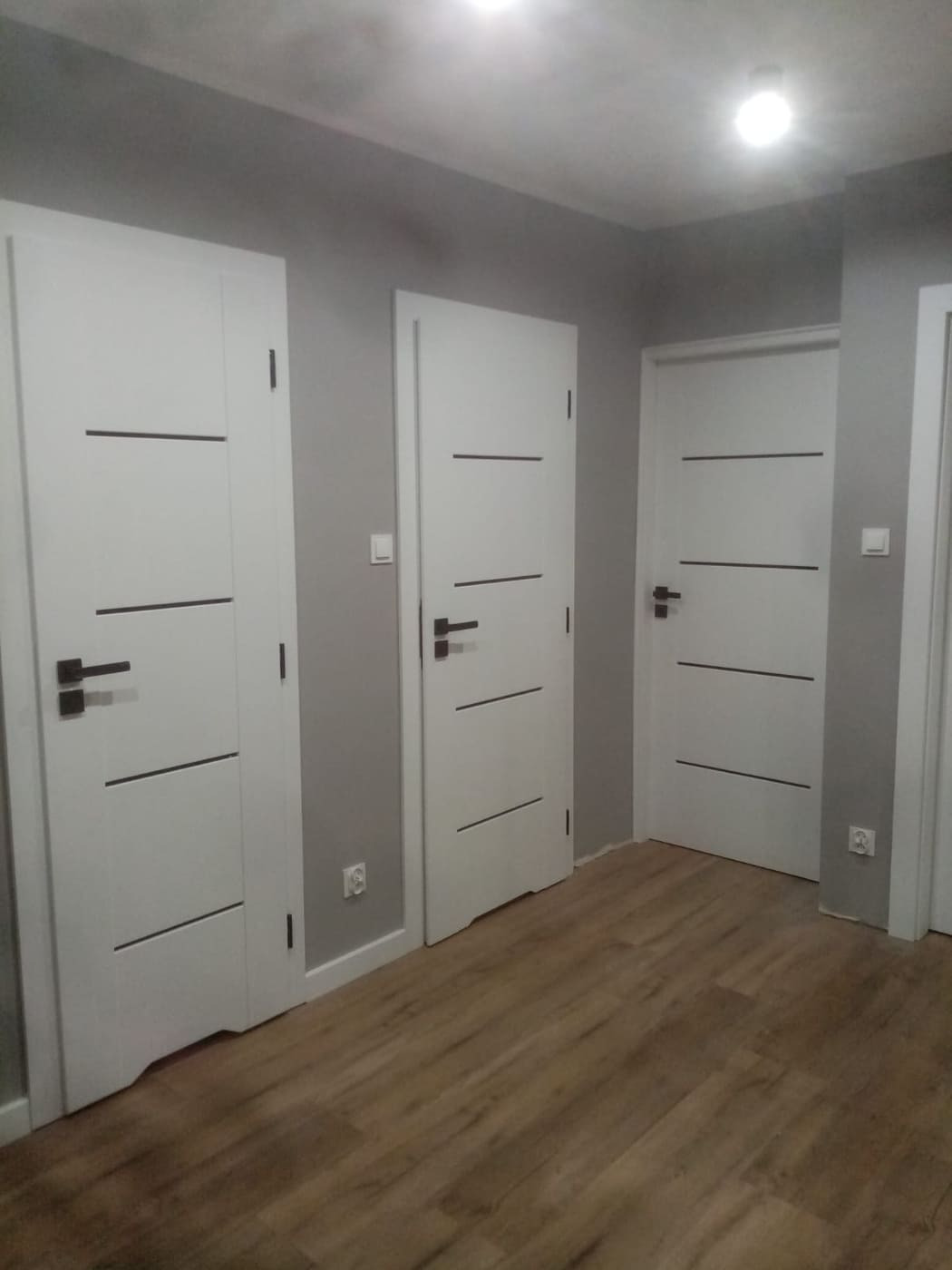 Trzy białe drzwi wewnętrzne z poziomymi, ciemnymi paskami i czarnymi klamkami, zamontowane w szarym pomieszczeniu z panelami podłogowymi w odcieniach brązu.