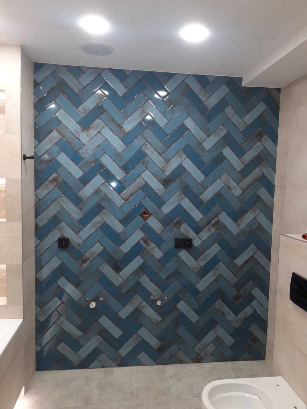 Ściana łazienki wyłożona błyszczącymi, niebieskimi płytkami w jodełkę, z widocznymi instalacjami hydraulicznymi i elektrycznymi przed montażem armatury.