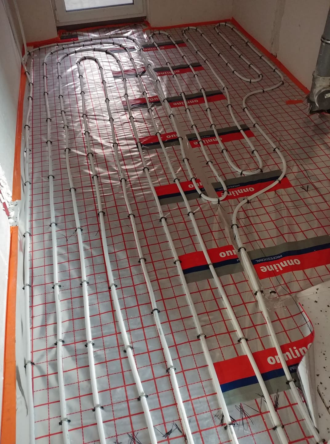 Rozłożone rury ogrzewania podłogowego na folii z siatką, widoczne uchwyty mocujące i fragment ściany z pomarańczowym zabezpieczeniem.