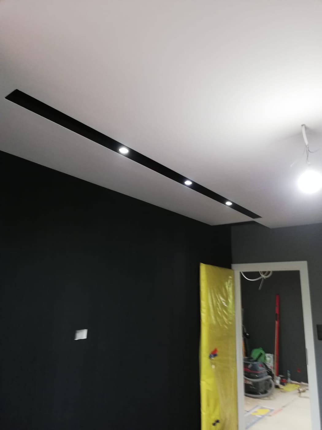 Czarne ściany i biały sufit z wpuszczonymi, liniowymi oprawami oświetleniowymi LED, widoczna folia ochronna na futrynie drzwi.