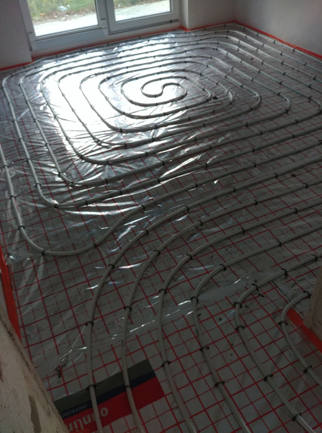 Rury ogrzewania podłogowego ułożone spiralnie na folii z czerwoną siatką, widoczne elementy mocujące rury i fragment ściany w surowym stanie.