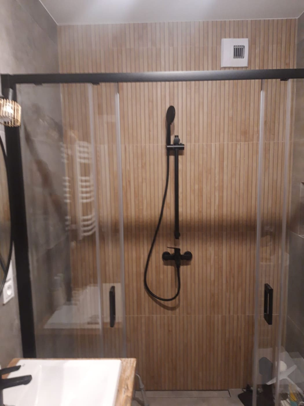 Nowoczesna kabina prysznicowa z czarną armaturą na tle płytek imitujących drewniane lamele, widoczny fragment umywalki z blatem.