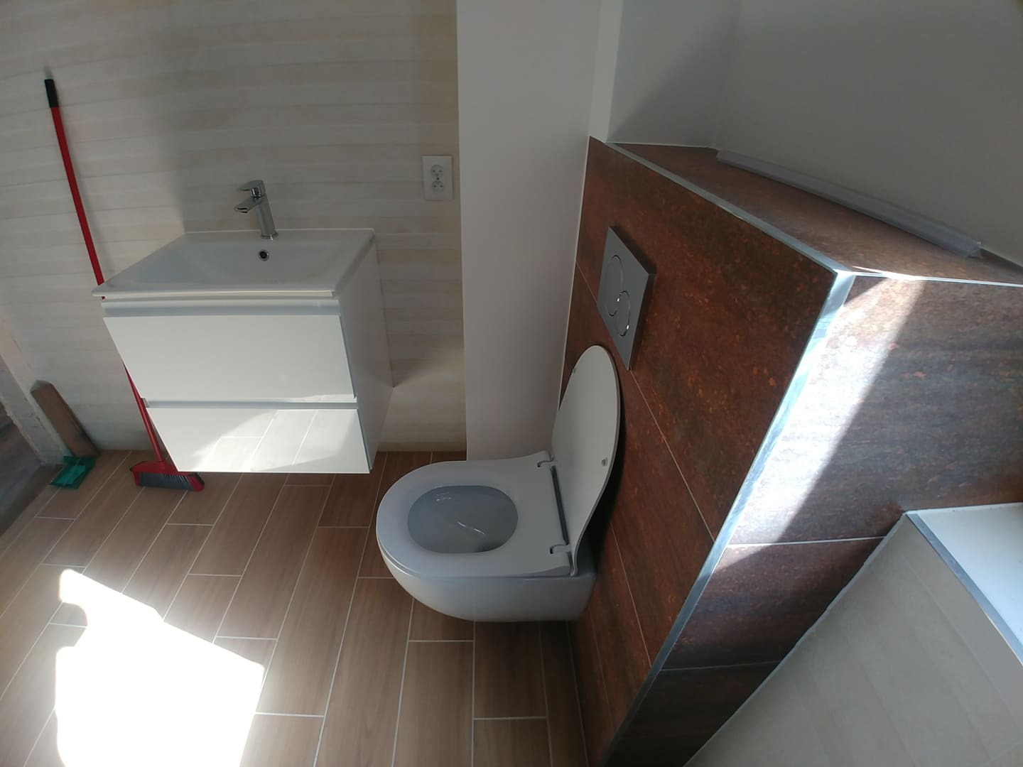 Nowoczesna łazienka z brązowymi płytkami imitującymi drewno, białą umywalką z szafką i podwieszaną toaletą z chromowanym przyciskiem spłukiwania, widoczna czerwona zmiotka.