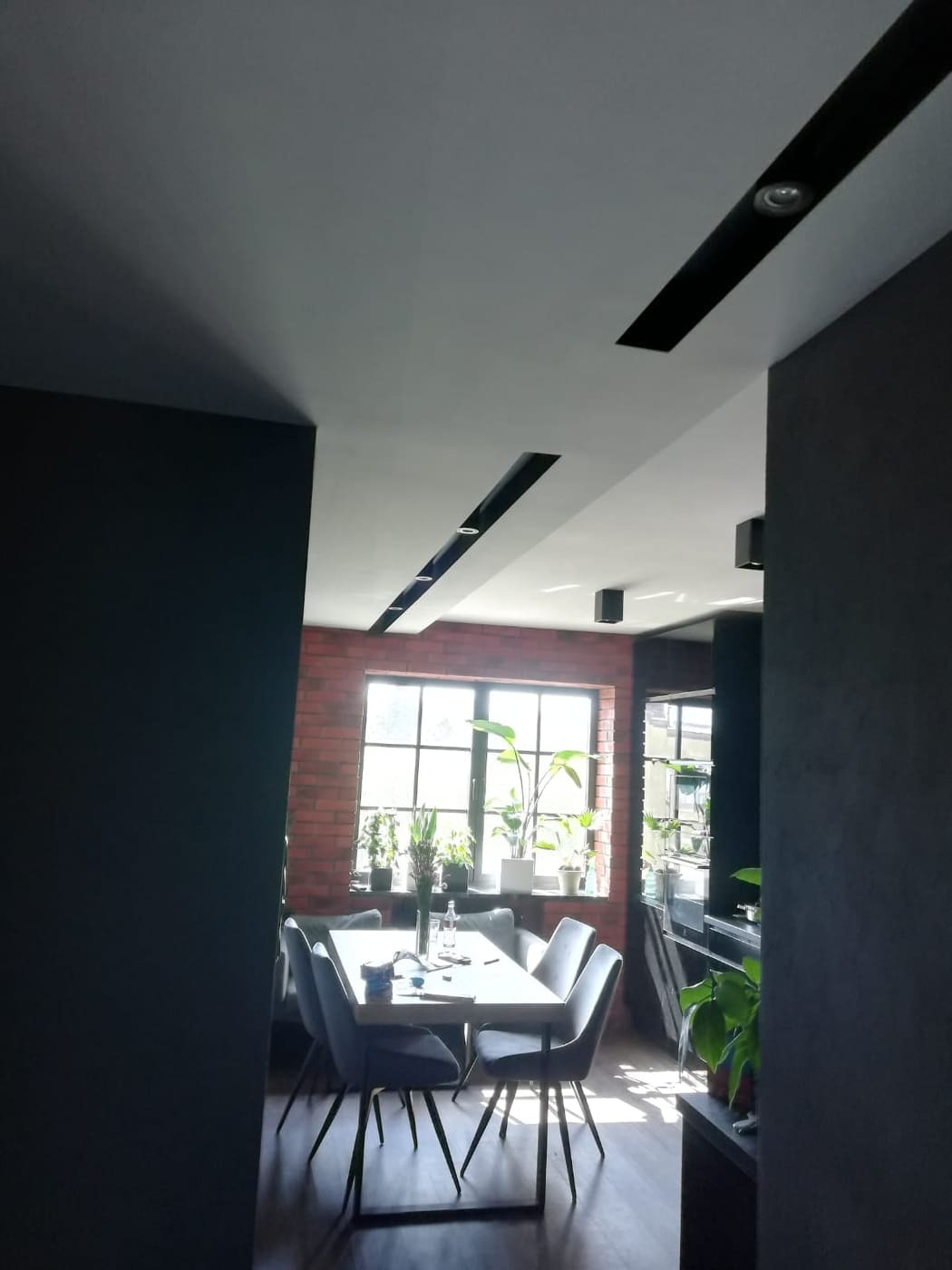 Nowoczesne wnętrze jadalni z ceglaną ścianą, białym stołem i szarymi krzesłami, oświetlenie sufitowe w czarnych listwach, widok z korytarza.
