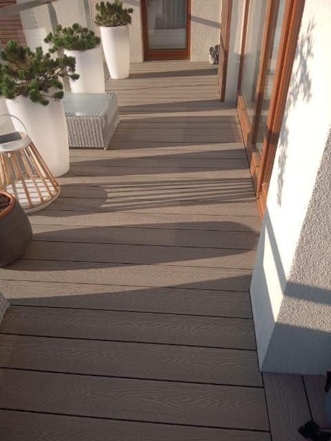 Wykończony taras z desek kompozytowych w odcieniach brązu, z białymi donicami z roślinami i rattanowym stolikiem, oświetlony popołudniowym słońcem rzucającym cienie.