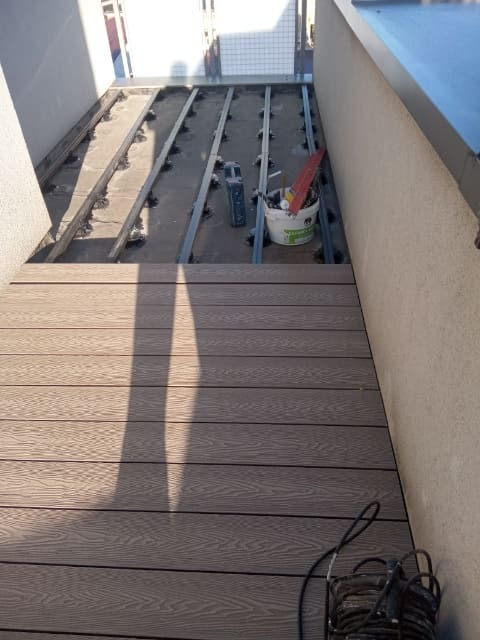 Balkon w trakcie montażu desek kompozytowych na legarach, widoczne narzędzia i wiadro z akcesoriami.