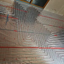 Rozłożona instalacja ogrzewania podłogowego z rurkami w kształcie meandrów na izolacji z nadrukowaną siatką, widoczne okno dachowe w tle.