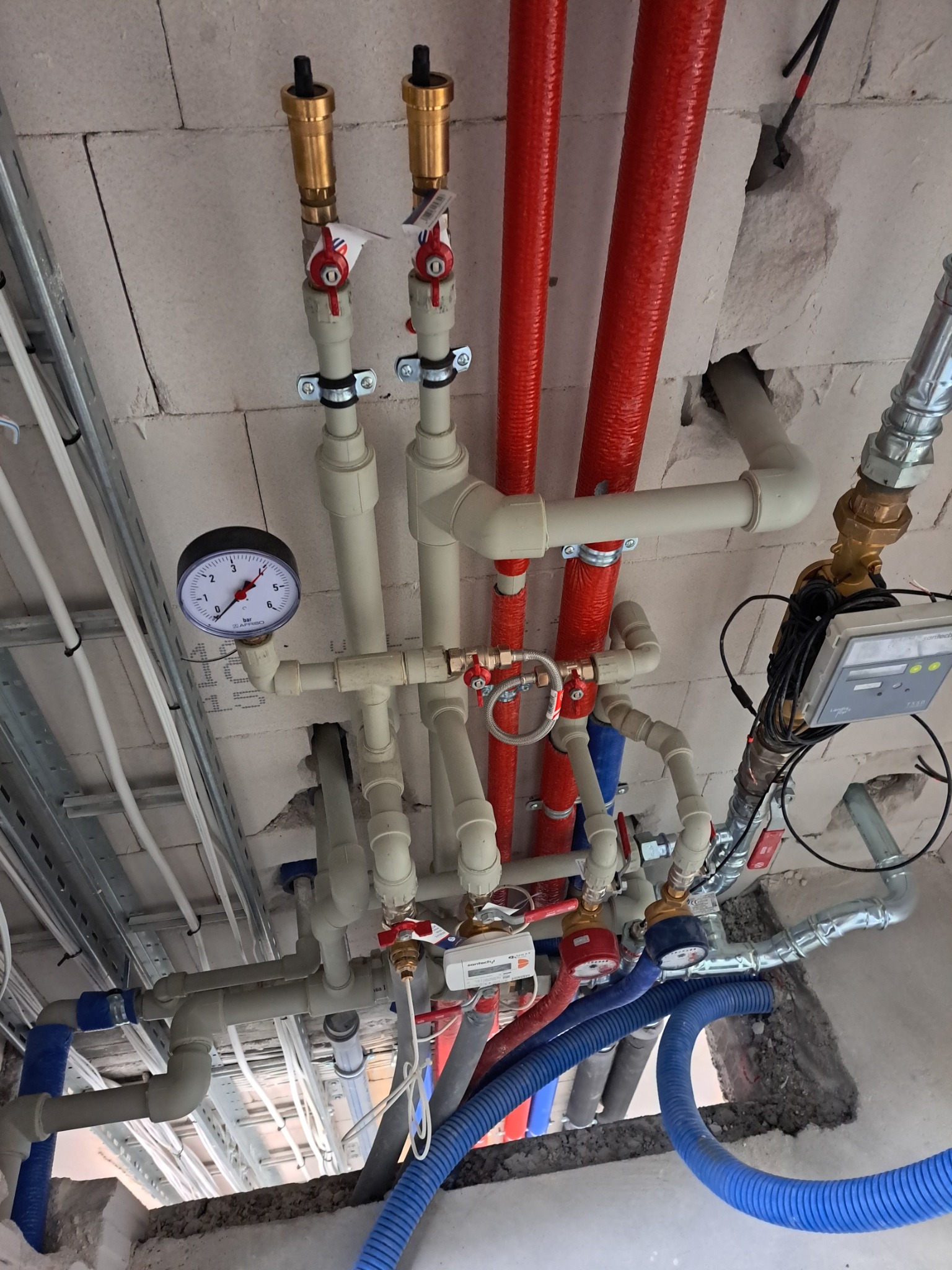 Widok od dołu na skomplikowaną instalację grzewczą i sanitarną pod stropem, z widocznymi rurami w kolorach czerwonym, niebieskim i białym, manometrem, zaworami i licznikiem ciepła.