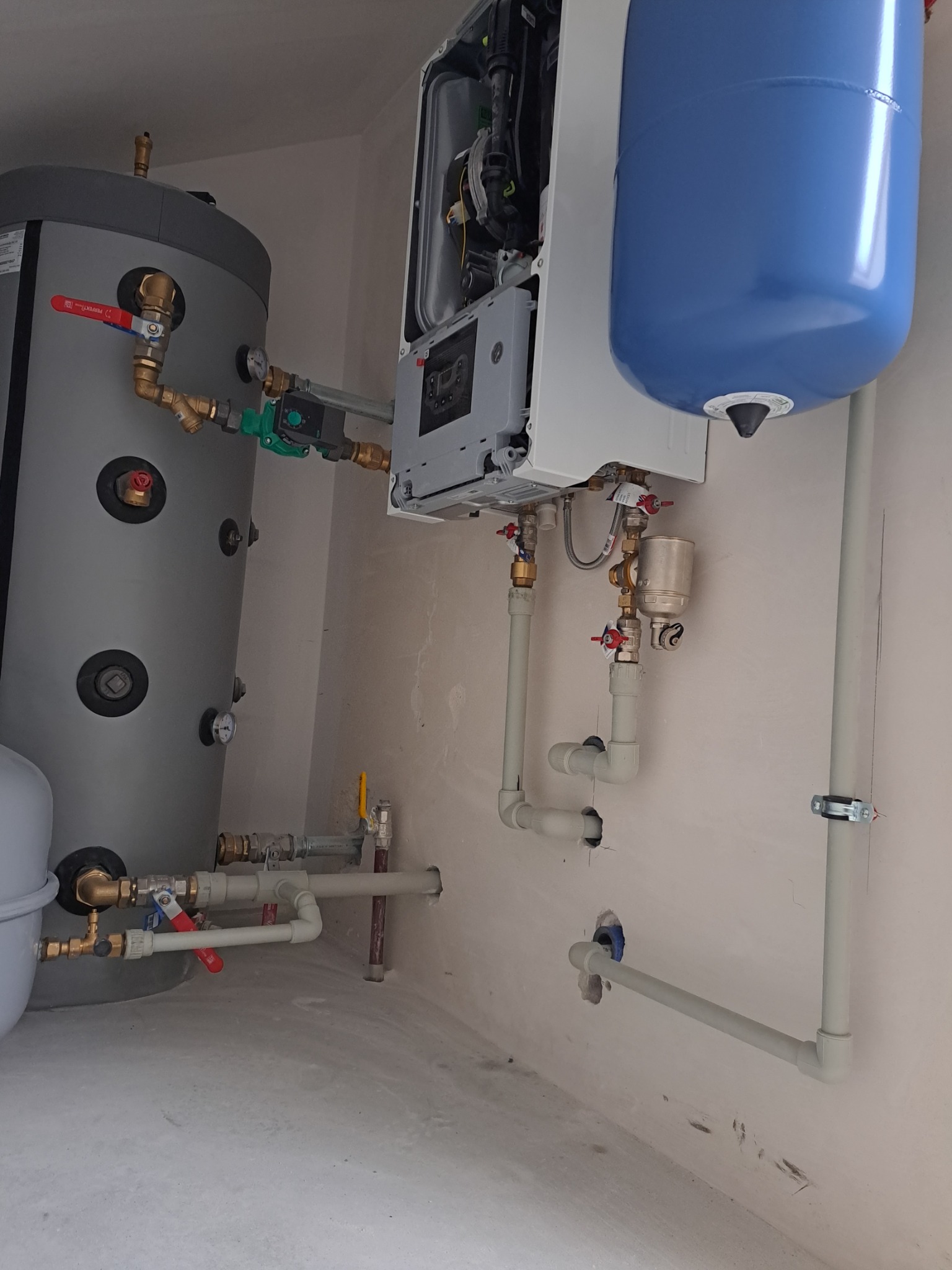 Szary zasobnik ciepłej wody użytkowej z czerwonymi zaworami, biały piec dwufunkcyjny z niebieskim naczyniem przeponowym i instalacja z szarych rur PCV w kotłowni.