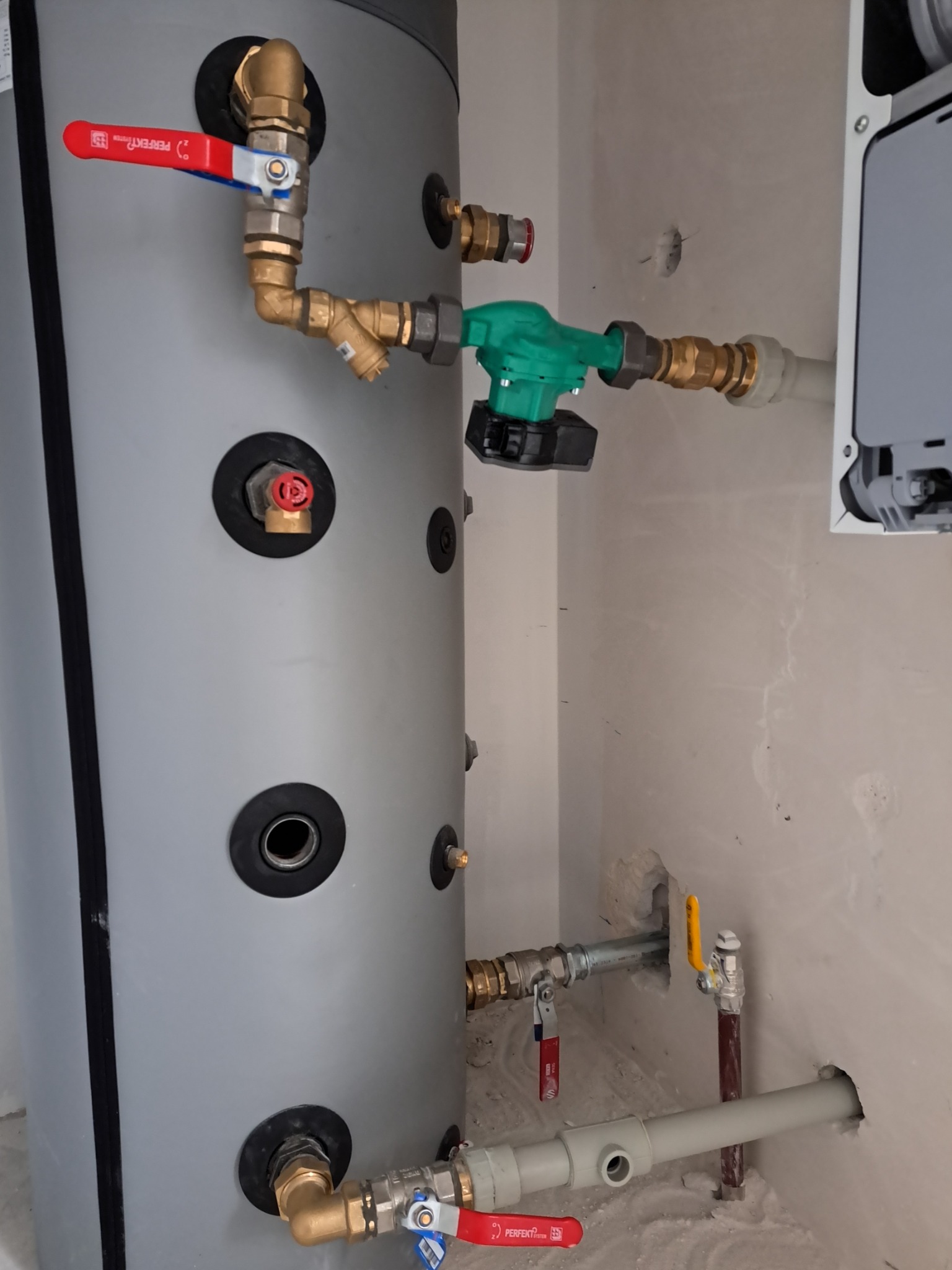 Szary zbiornik buforowy z licznymi przyłączami hydraulicznymi i zaworami kulowymi z czerwonymi rączkami, zainstalowany w pomieszczeniu w Wołominie, widoczne rury miedziane i plastikowe.