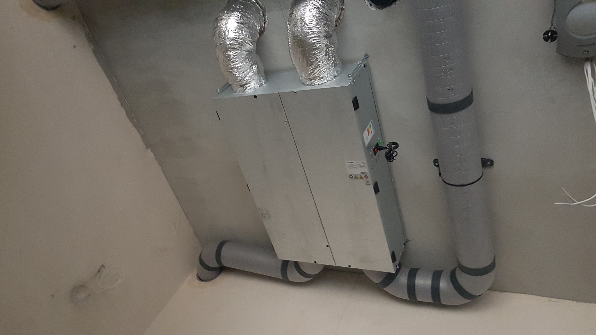 Centrala wentylacyjna z rekuperacją, widoczne rury wentylacyjne spiro i PCV, zamontowana pod sufitem w surowym pomieszczeniu.