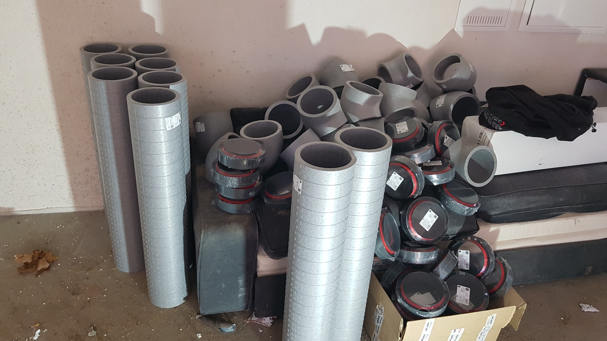 Stos rur wentylacyjnych z szarego tworzywa, kolanek i elementów montażowych w magazynie, niektóre jeszcze w opakowaniach.