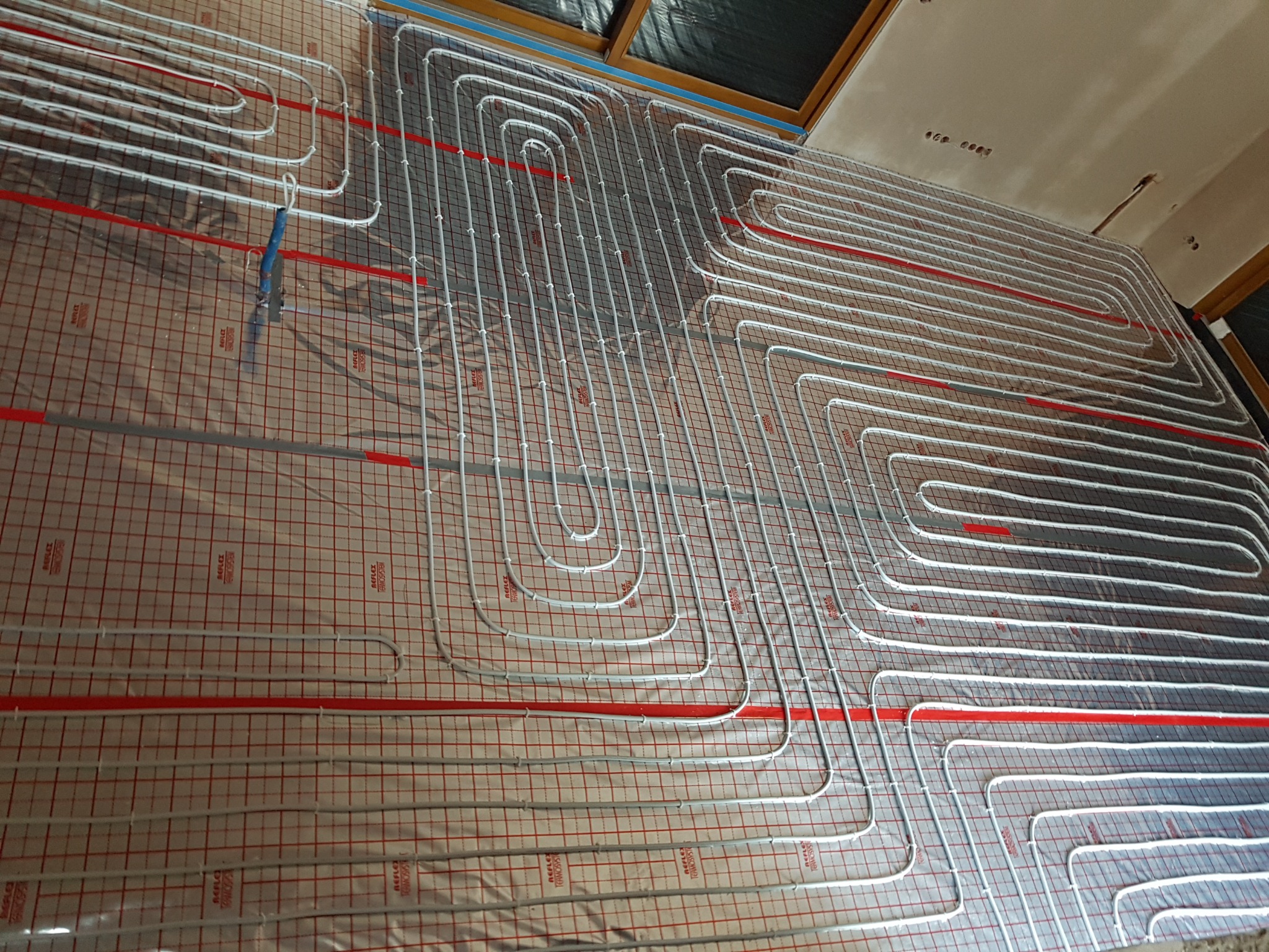 Rozłożona instalacja ogrzewania podłogowego z rurkami w kształcie meandrów na izolacji z nadrukowaną siatką, widoczne okno dachowe w tle.