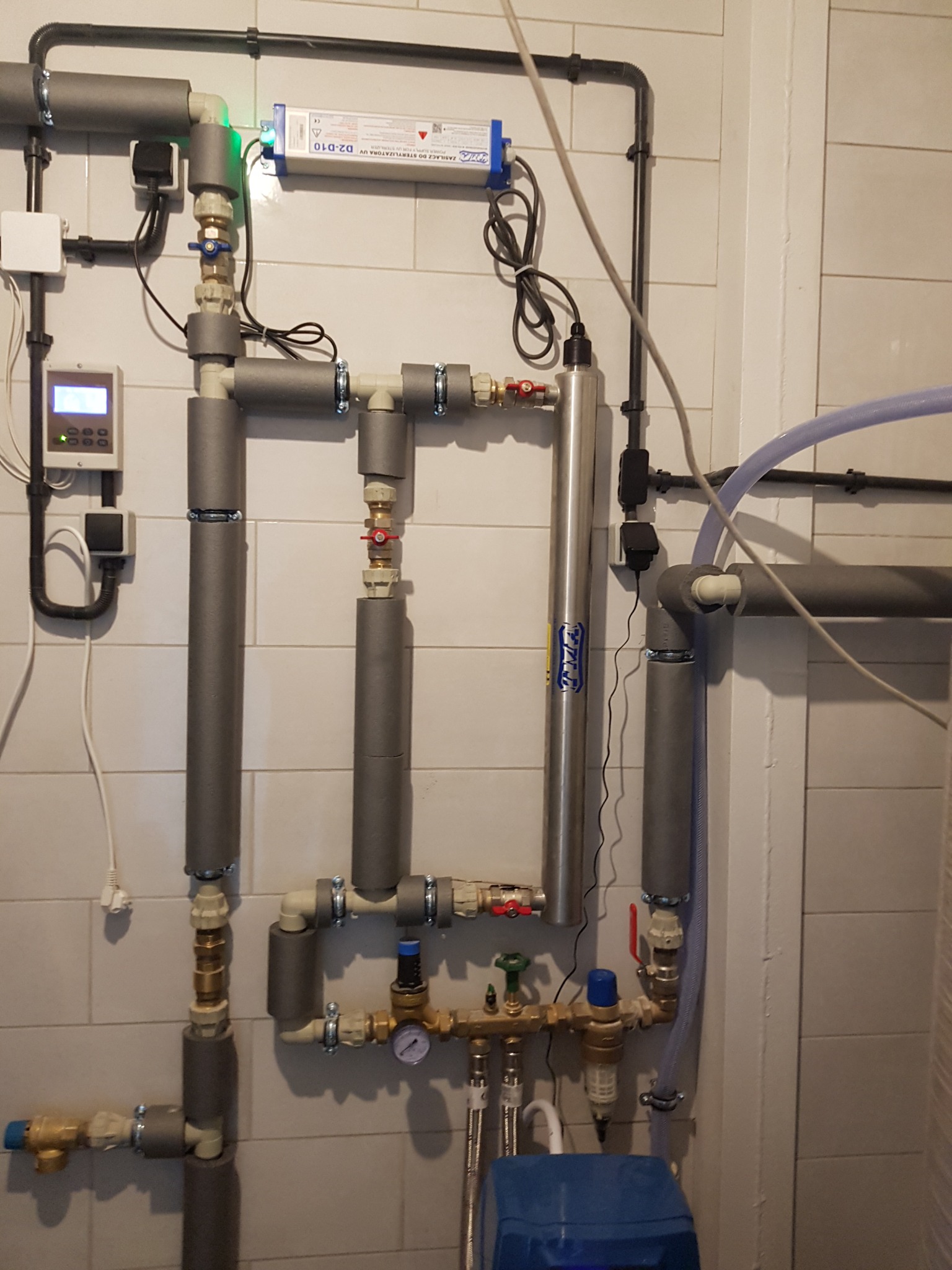 Złożony system rur z izolacją termiczną, zaworami i manometrem na tle białych płytek, widoczny również sterylizator UV wody oraz niebieski filtr mechaniczny.