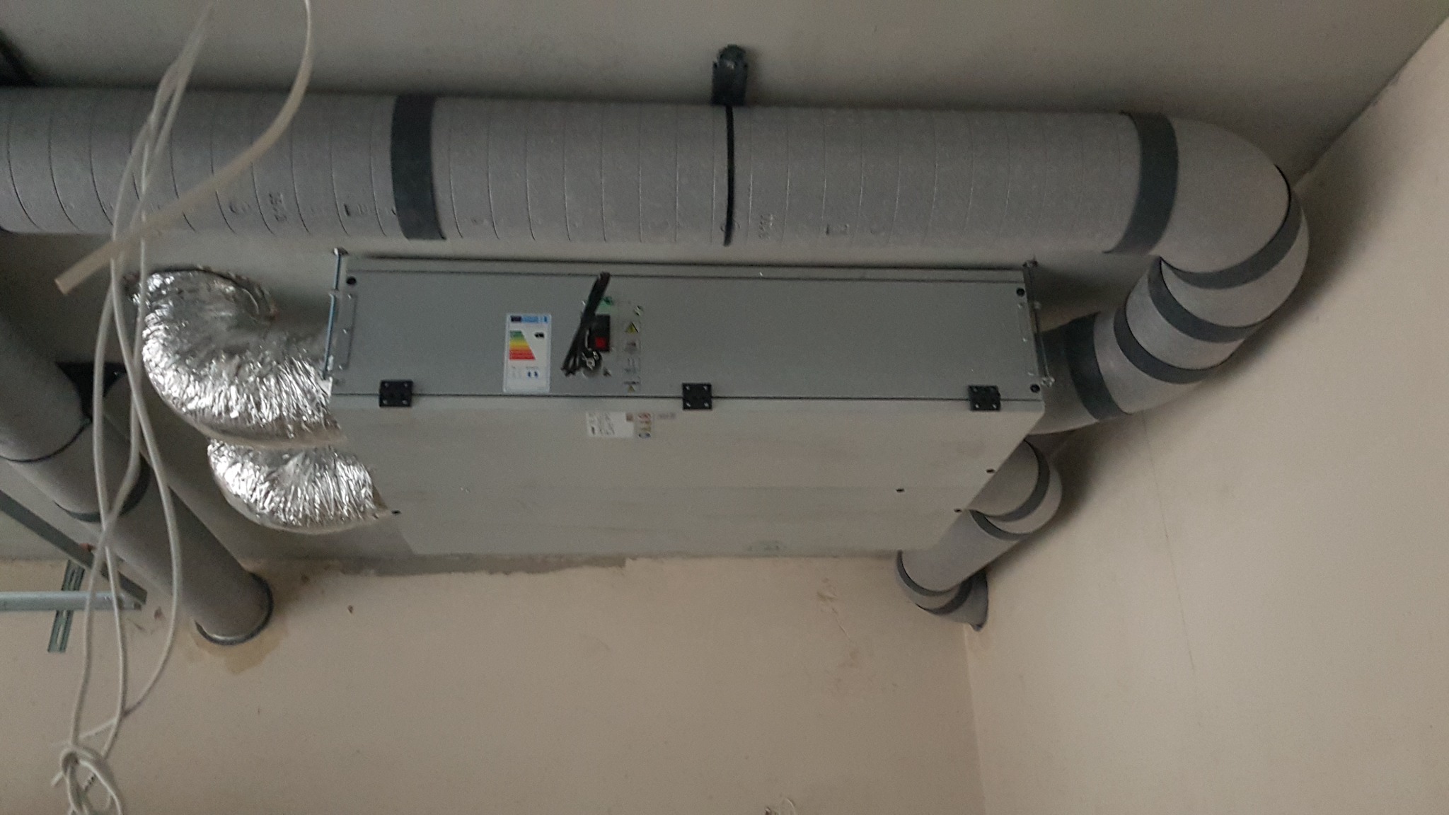 Centrala wentylacyjna z podłączonymi przewodami wentylacyjnymi, widoczna etykieta energetyczna i elementy montażowe na suficie w trakcie instalacji.