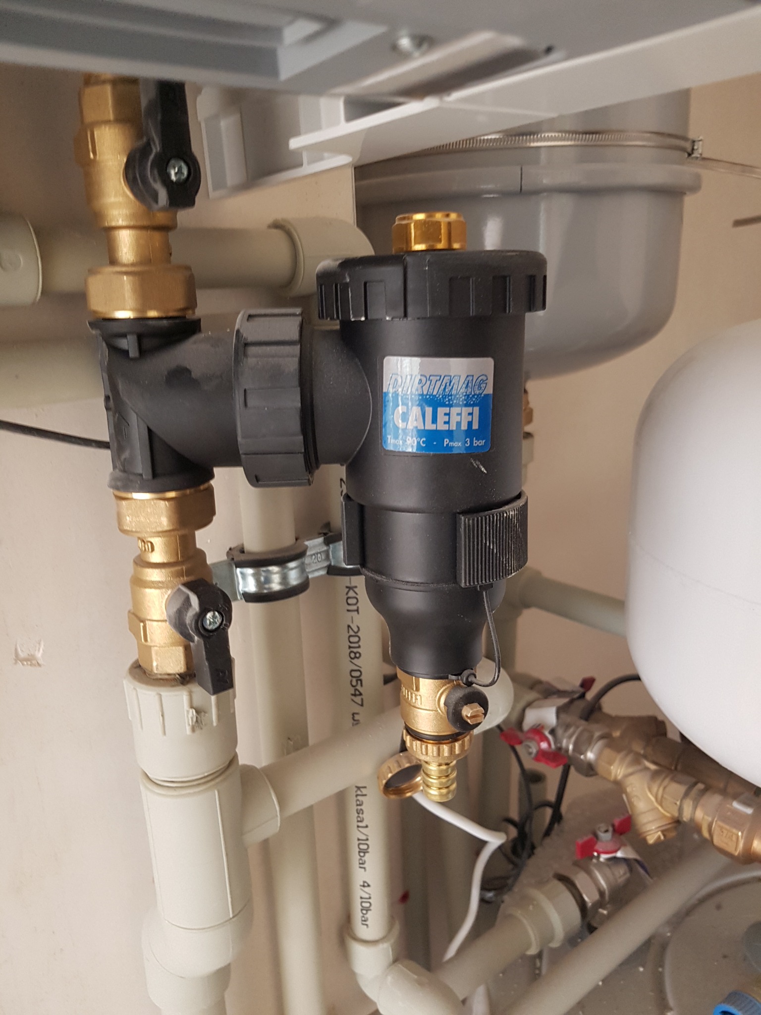 Zamknięty filtr magnetyczny Caleffi Dirtmag zamontowany na instalacji centralnego ogrzewania, widoczne zawory kulowe z czarnymi motylkami, rury z tworzywa sztucznego i zbiornik wyrównawczy.