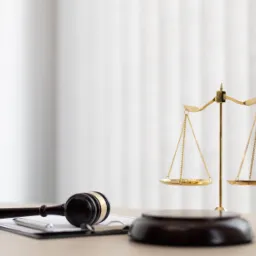 Złota waga sprawiedliwości i drewniany młotek sędziowski na biurku z dokumentami i długopisem, na tle białych zasłon.
