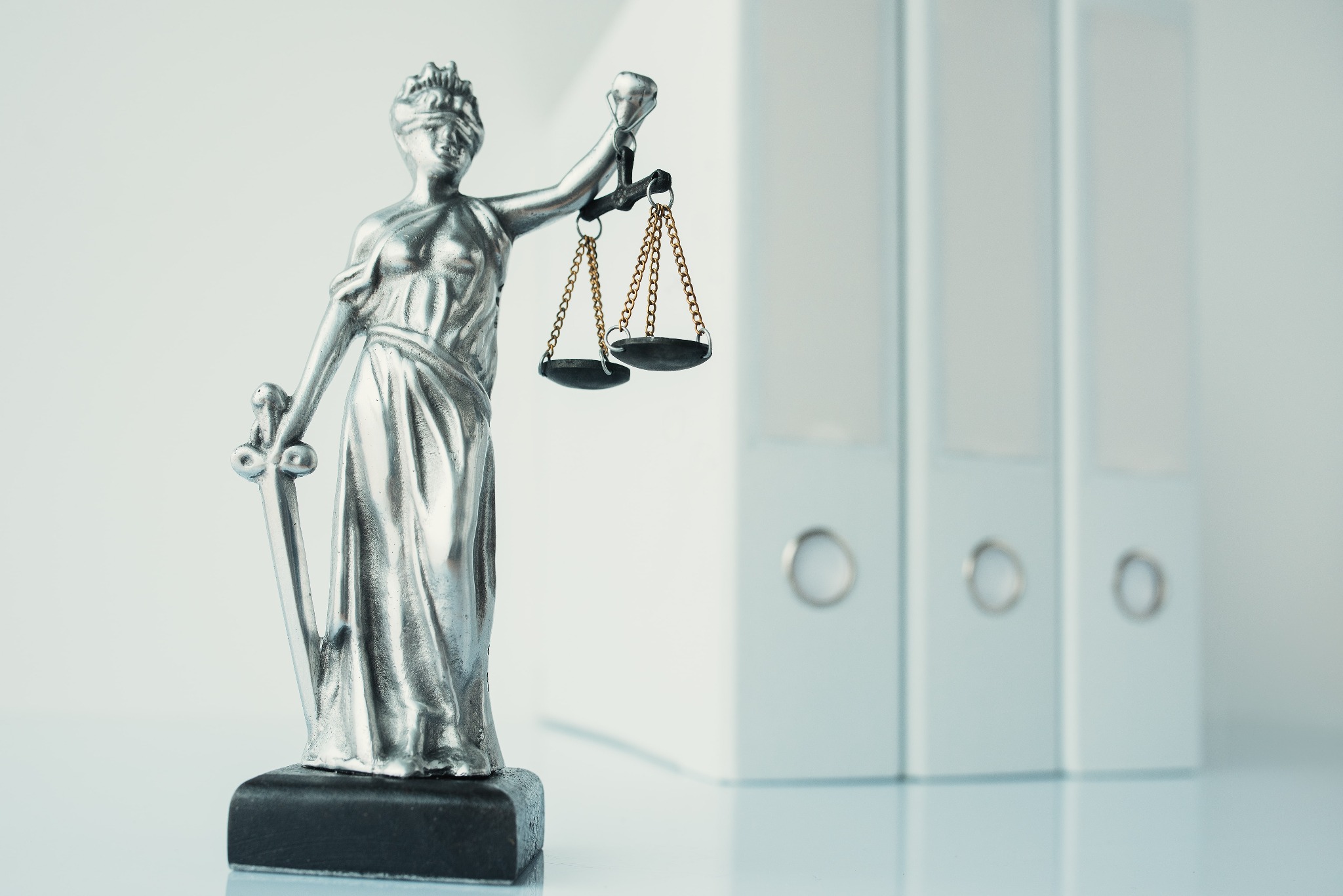 Statuetka Temidy ze srebrnego metalu z wagą i mieczem na tle trzech białych segregatorów.