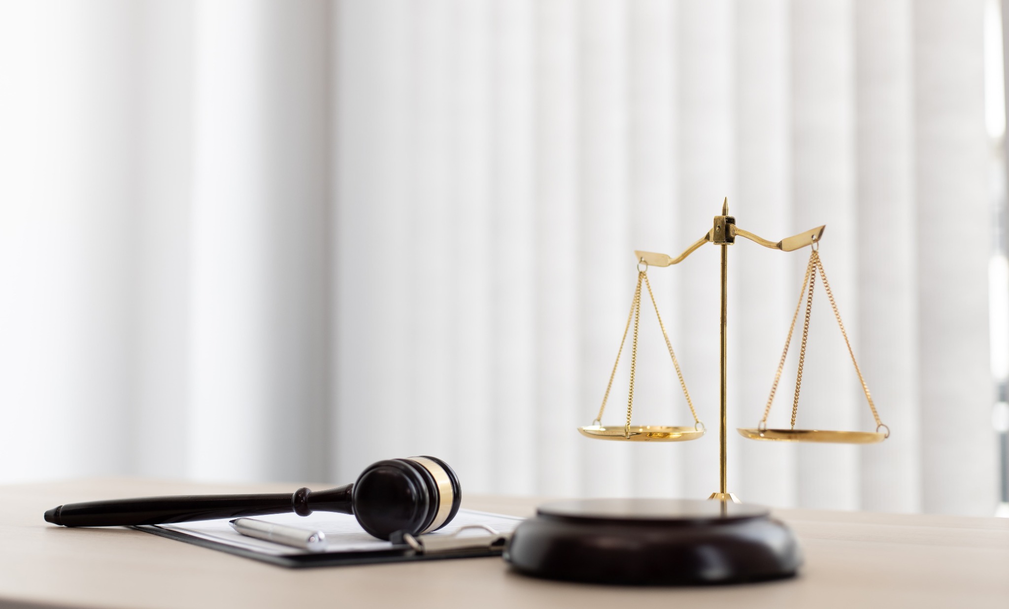 Złota waga sprawiedliwości i drewniany młotek sędziowski na biurku z dokumentami i długopisem, na tle białych zasłon.