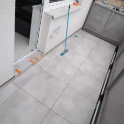 Wykończony balkon z szarymi płytkami, niebieski mop oparty o ścianę, widoczne zabezpieczenia z taśmy malarskiej przy progu drzwi balkonowych.