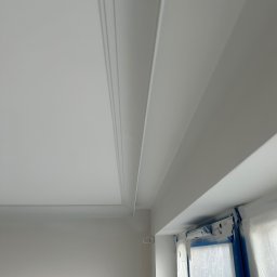 Olaf Nowicki - Białe, świeżo pomalowane wnętrze z dekoracyjną listwą sufitową i zabezpieczonym oknem folią malarską. Widok z dołu, podkreślający detale wykończenia.