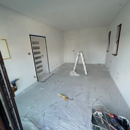 Olaf Nowicki - Białe wnętrze po malowaniu, zabezpieczone folią, z drabiną i narzędziami malarskimi. Widoczne świeżo pomalowane ściany i marmurowe płytki podłogowe.