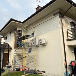 Olaf Nowicki - Dom w trakcie malowania elewacji, widoczne rusztowanie, częściowo pomalowana ściana z drewnianą okładziną i zabezpieczone okna folią malarską.