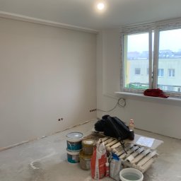 Olaf Nowicki - Wnętrze pokoju w trakcie malowania: świeżo pomalowane ściany w odcieniach bieli, widoczne puszki z farbą, narzędzia malarskie i grzejnik przygotowany do malowania.