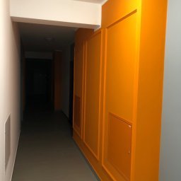 Olaf Nowicki - Korytarz po remoncie: pomarańczowa ściana z panelami i drzwiczkami rewizyjnymi, szara ściana i podłoga, biały sufit. Nowoczesne wykończenie wnętrza.