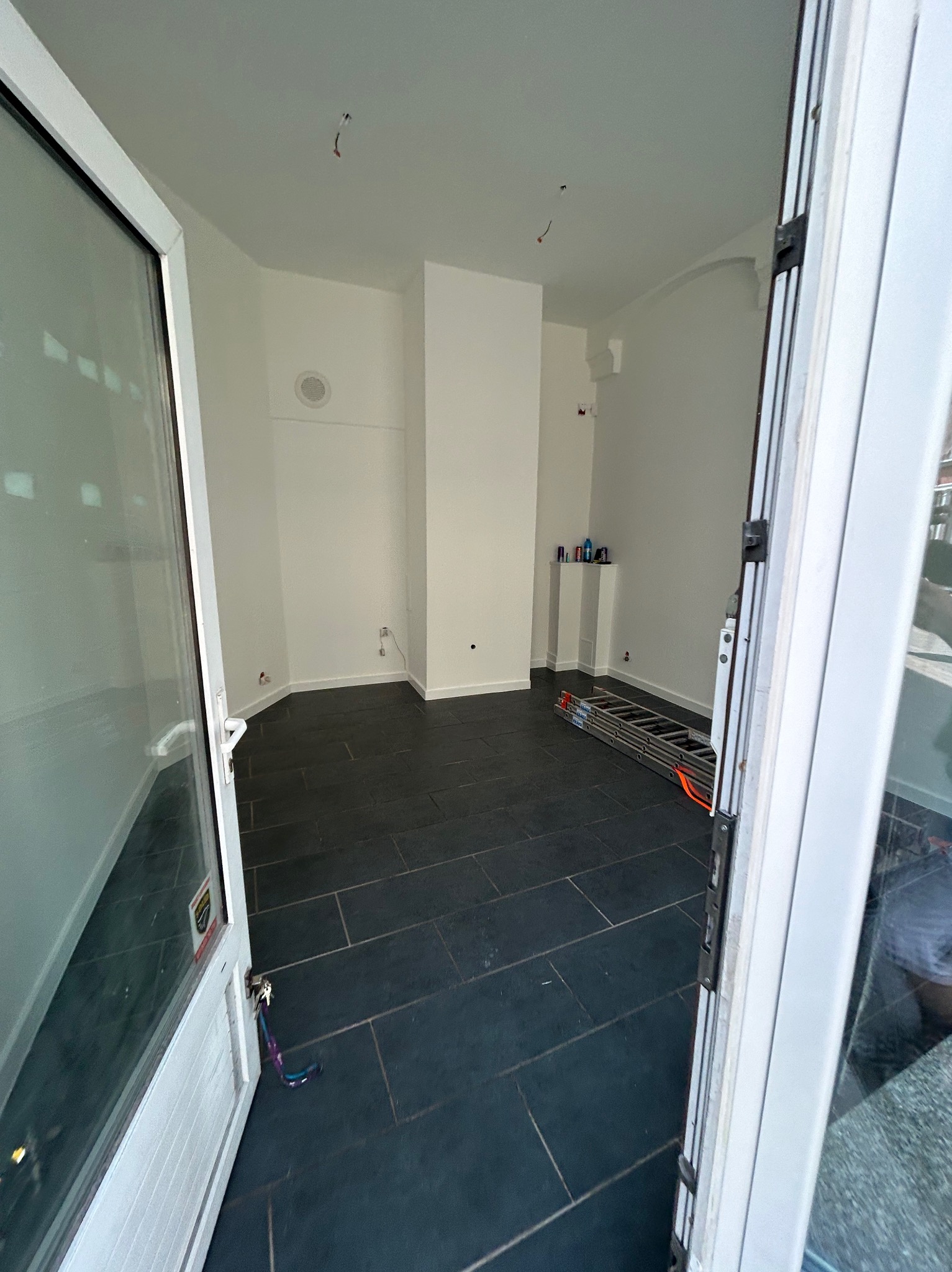 Puste, jasne wnętrze z ciemną podłogą z płytek, widok przez otwarte białe drzwi balkonowe. W tle drabina oparta o ścianę i białe ściany z wystającymi kablami.