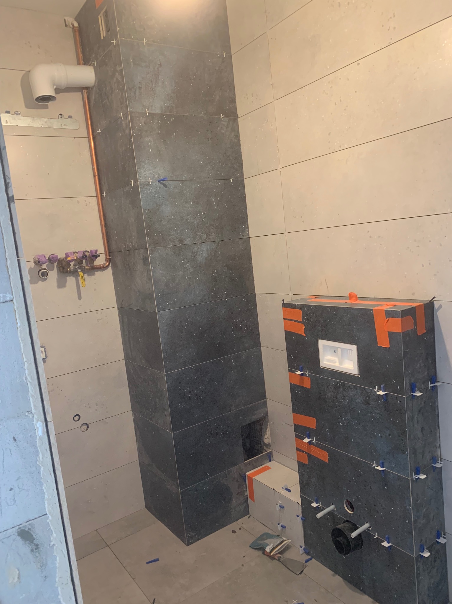 Montaż ciemnych, nakrapianych płytek w łazience, widoczne rury i elementy instalacji. Etap prac wykończeniowych, z użyciem krzyżyków glazurniczych.