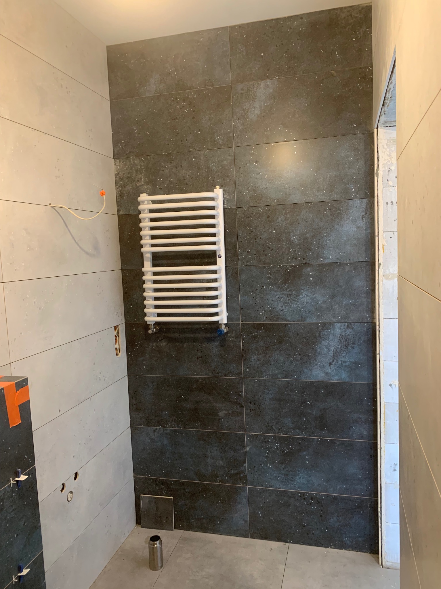 Łazienka w trakcie wykańczania: ułożone szare płytki na ścianach, biały grzejnik drabinkowy, widoczne elementy instalacji. Minimalistyczny design, surowy klimat.